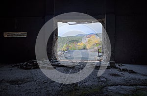 View through the door of abandoned factory with rails and broken brick wall inside and autumn landscape aoutside
