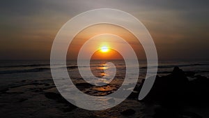 A view from a drone flying over the sea with waves and a view of the fiery sunset above the water with rocks below