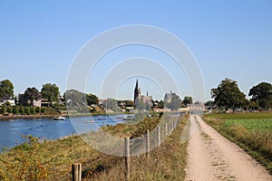 View on dutch cycling track along river Maas with village and church background in autumn on sunny day - Kessel, Netherlands