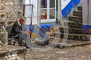 View of elderly with typical coat, sitting on wooden bench, with rain hat open on the ground, in the square of the medieval