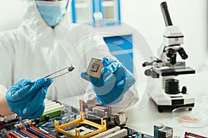 View of engineer holding microchip and small stone near microscope and computer motherboard