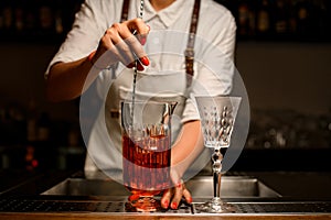 view of female bartender gently stirring with spoon cocktail in transparent mixing cup
