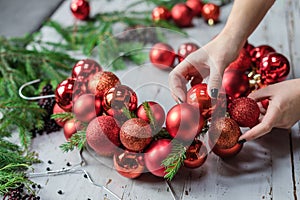 View of female hands making Christmas wreath with fir branches and decorative red balls on wooden rustic tabletop