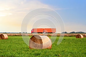 View of a field with hay in rolls against the background of trucks with semi-trailer driving along the highway. Harvesting dry