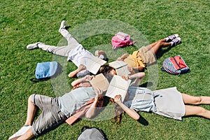 View of four multicultural schoolkids lying on lawn near backpacks and covering faces with books