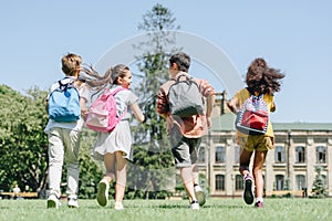 View of four multiethnic schoolkids with backpacks running on lawn in park