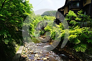 View of fresh stream with stone bank through green trees and local buildings on stone bank in Kurokawa onsen town