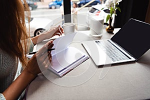 View of girl studying online with notepad and laptop in cafe with cup of coffee