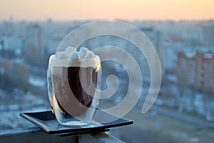 A view of a glass mug with hot black coffee and marshmallows against the background of the dawn sky and the city skyline