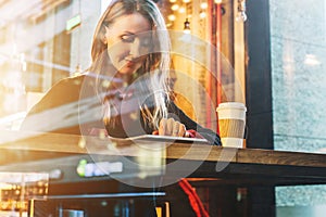 View through glass. Young smiling business woman sitting in cafe at table, drinking coffee and using tablet computer.