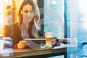 View through glass. Young smiling business woman sitting in cafe at table, drinking coffee and using tablet computer.