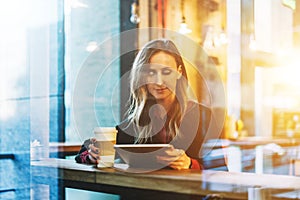 View through glass. Young smiling business woman sitting in cafe at table, drinking coffee and using tablet computer.