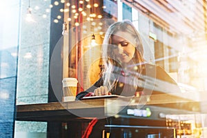 View through glass. Young smiling business woman sitting in cafe at table, drinking coffee and using tablet computer.