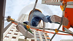 View from the ground on kid in sneakers standing on high tower at playground and walkng over rope bridge