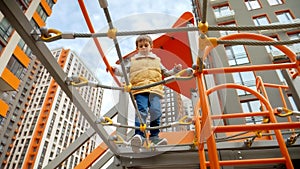 View from the ground on little boy walking over the rope bridge at new public playground. Active child, sports and development,