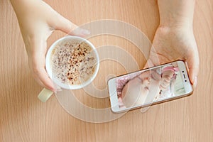 View handheld color video baby monitor. Female hands are holding a smartphone with a baby monitor app. Near hot drink