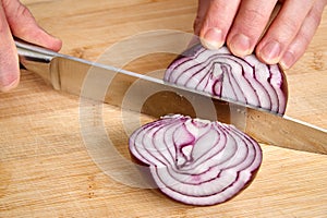 View on hands of a man chopping peeled fresh red onion for lunch an a bamboo cutting board.