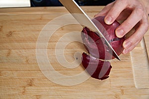 View on hands of a man chopping and peeling a fresh red onion for lunch an a bamboo cutting board.