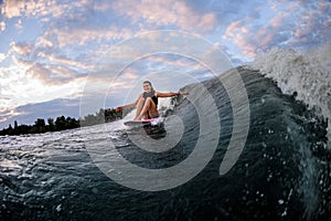 View on happy woman riding wave while sitting on surf style wakeboard with arms outstretched to the sides