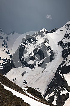 View of a high steep rocky mountain partially covered with snow against a dark cloudy sky with clouds clinging to the