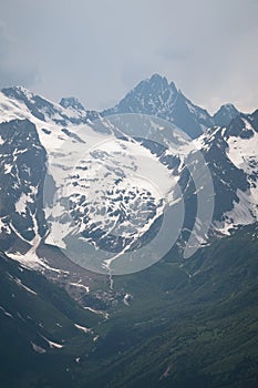 View of a high steep rocky mountain partially covered with snow against a dark cloudy sky with clouds clinging to the