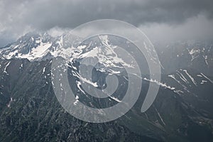 View of a high steep rocky mountain partially covered with snow against a dark cloudy sky with clouds clinging to the
