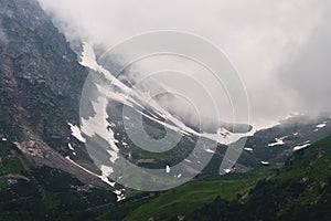 View of a high steep rocky mountain partially covered with snow against a dark cloudy sky with clouds clinging to the