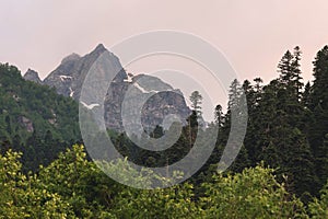 View of a high steep rocky mountain partially covered with snow against a dark cloudy sky with clouds clinging to the