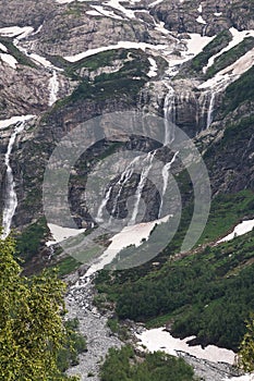 View of a high steep rocky mountain partially covered with snow and waterfalls against a dark cloudy sky with clouds