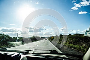 View of a highway full of greenery on both sides inside a car and with the strong sun and blue sky.