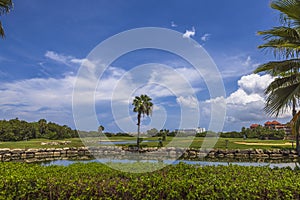 View of hotel building and blue water surface of pond and green grass golf field on background blue sky with white clouds.