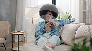 View through house plant happy smiling relaxing African American woman carefree girl resting on couch at home scrolling