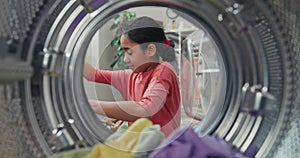 A view from inside the drum into the laundry room. A cute little girl is putting colorful clothes in the wash. A child