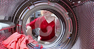 View from inside the washing machine drum, a young woman loading dirty laundry from a basket. Taking clothes out of washing