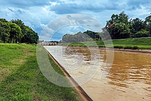 View of irrigation canal with weeds and debris flowing together with strong flowing dirty brown water due to soil and mud from photo