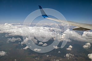 View of jet airplane wing from inside flying over white puffy clouds in blue sky. Travel and air transportation concept