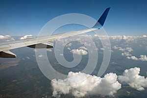 View of jet airplane wing from inside flying over white puffy clouds in blue sky. Travel and air transportation concept