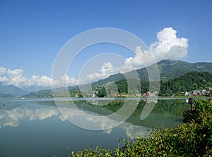 View of the lake with turquoise water and the opposite shore near the mountain Sarangkot. Reflection of clouds in calm water.