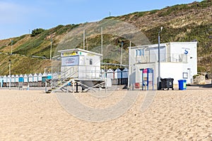 A view of a lifeguard tower on a sandy beach with beach huts and grassy cliff in the background under a beautiful blue sky