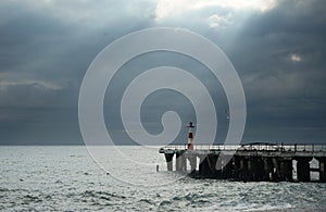 View of the lighthouse on the stone pier near the stormy sea with sun rays through the dark sky