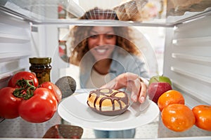 View Looking Out From Inside Of Refrigerator As Woman Opens Door And Reaches For Unhealthy Donut