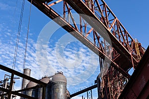 View looking up through rusting metal structures of a steel manufacturing plant, blast furnaces behind, blue sky with light clouds