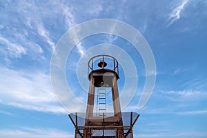 View looking up at a weathered steel light house tower at the end of a pier with blue sky and bright white feathery clouds in the