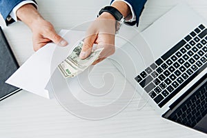 View of man holding envelope with money near laptop on table