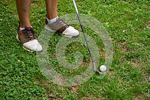 View of man`s feet with golf shoes with iron and cue ball, ready to hit, on green grass