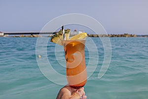 View of man's hand holding a glass of alcoholic cocktail on background of blue water of Caribbean Sea.