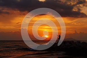 View of a man silhouette standing on sea stone beach and shooting selfie with beautiful amazing sunset of orange red colors. Beaut