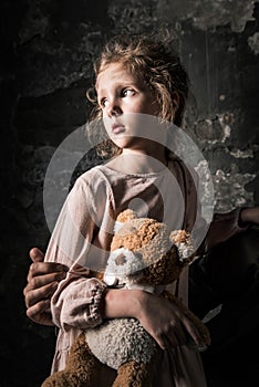 View of man touching sad kid with teddy bear in dirty room, post apocalyptic concept