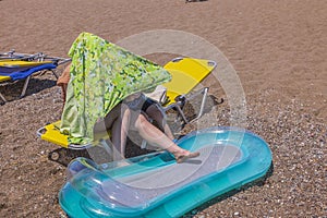 View of mother and child on sun lounger on sandy beach with air mattress for swimming, hiding from sun under towel.