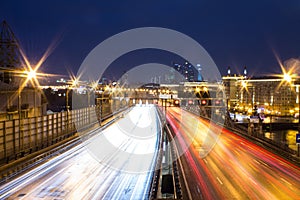 View of multilane urban highway with glowing lines of light from car headlamps and modern architecture